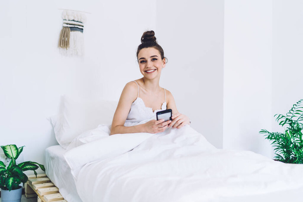 Content brunette waking up on comfortable soft bed with white blanket using smartphone in room with trendy minimalist interior and looking at camera - Photo, Image