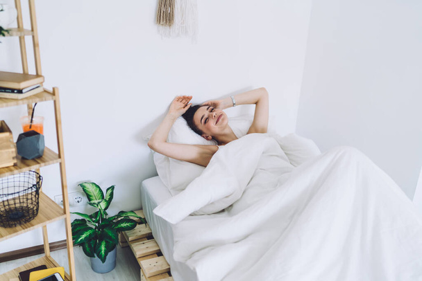 Happy young woman waking up fully rested after good sleep and enjoying morning while lying with raised hands in single bed in light room - Photo, image