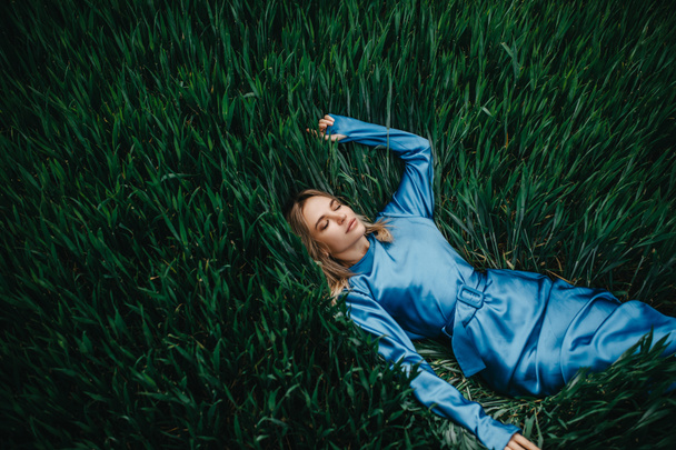 A mulher jovem em um vestido azul encontra-se, relaxa e descansa entre o trigo verde. Vista superior. Espaço de cópia
. - Foto, Imagem