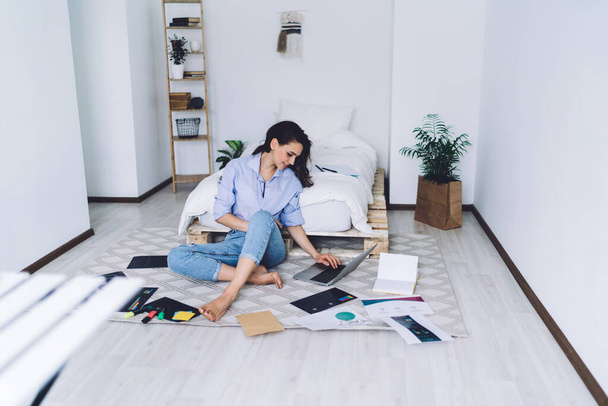 Female in casual clothes siting on floor in light stylish bedroom among highlighters and charts on paper while using laptop - Фото, изображение