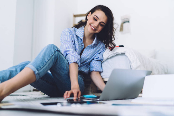 Side view of satisfied smiling woman in casual outfit typing laptop while sitting barefoot on floor with charts and papers in modern apartment - 写真・画像