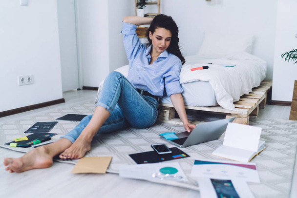 Thoughtful serious woman in casual outfit browsing laptop while sitting barefoot on floor with graphs and notes in modern room - Foto, afbeelding