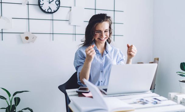Pretty adult woman in blue shirt and glasses with raised arms concentrating on laptop screen while sitting at desk against white wall with black decor and clock - Photo, image