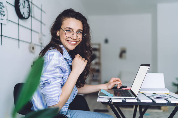 Side view of pretty cheerful woman in glasses and blue shirt sitting at desk browsing laptop with white screen and looking at camera - Foto, Imagen