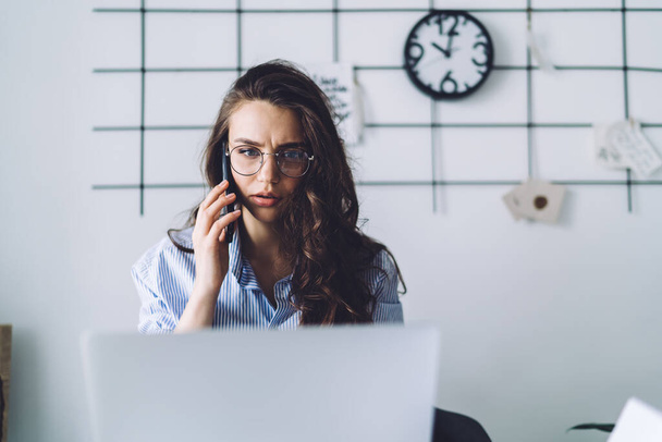 Anxious female worker with wrinkled nose bridge in glasses concentrating on multitasking speaking on smartphone and using laptop in office - Photo, image