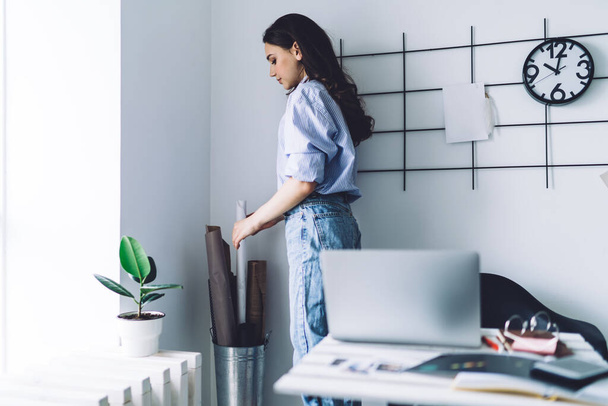 Side view of confident young woman in blue jeans and blouse standing in corner of office room by wall and taking white roll of paper - Foto, imagen