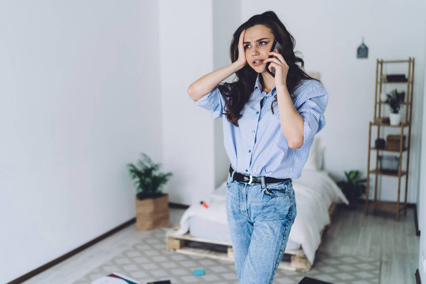 Eventful gloomy black haired female in casual jeans and shirt talking on mobile phone with hand on head in sunny bedroom and looking away - Foto, Imagem