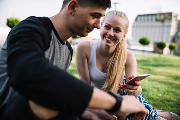Nevető fiatal nő mutatja vicces videó a mobiltelefon férfi barátja, miközben pihen együtt zöld gyep a városi parkban nyáron nap - Fotó, kép
