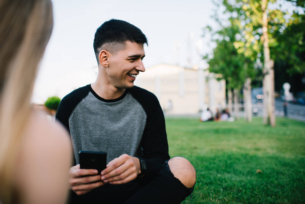 Satisfied optimistic carefree guy looking away while browsing modern mobile phone during sitting with friend on grass of park in summer - Foto, Bild