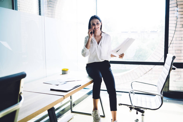Happy female entrepreneur having conversation with business partner on smartphone and reviewing notes while sitting on table in bright room and looking at camera - Foto, Imagen