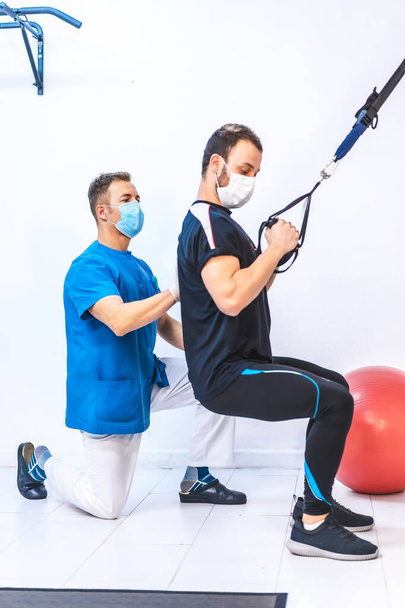 Physiotherapist in blue gown and a patient in a squat working the arms with rubber bands. Physiotherapy with protective measures for the Coronavirus pandemic, COVID-19. Osteopathy, sports chiromassage - Photo, image