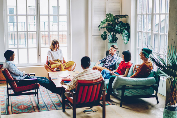 Grupo de personas relajadas y diversas sentadas en cómodos asientos alrededor de la mesa y comunicándose entre sí durante la reunión en la elegante sala de estar - Foto, Imagen