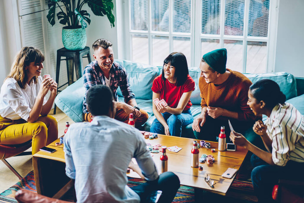 From above joyful immersed diverse hipsters playing cards with chips for poker around table and having fun in modern apartment - 写真・画像