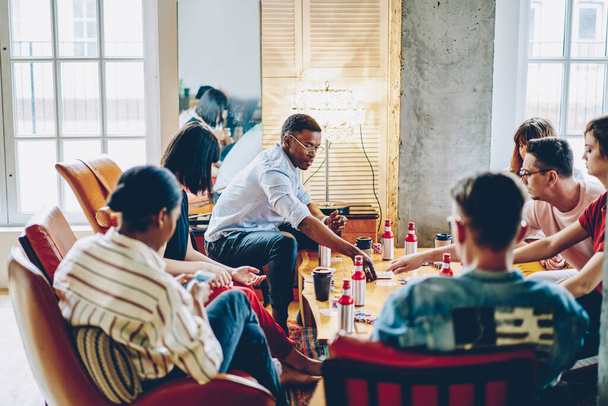 Group of focused diverse young people wearing casual clothes playing cards while sitting at table with beverages in loft apartment  - Fotoğraf, Görsel