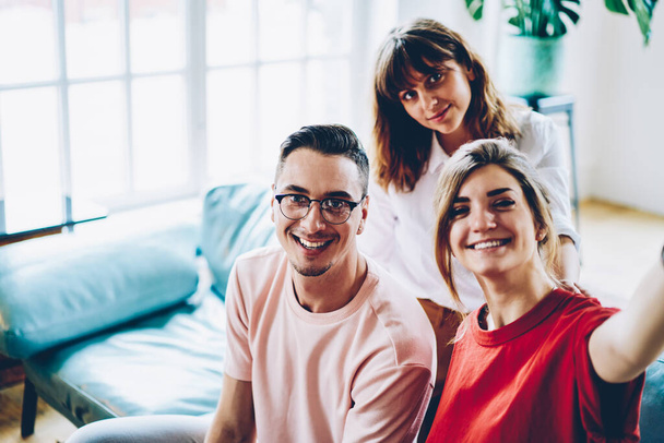 Smiling cheerful group of young smart friends in casual apparel making selfie sitting on sofa in cozy room with plant in pot  - Фото, изображение