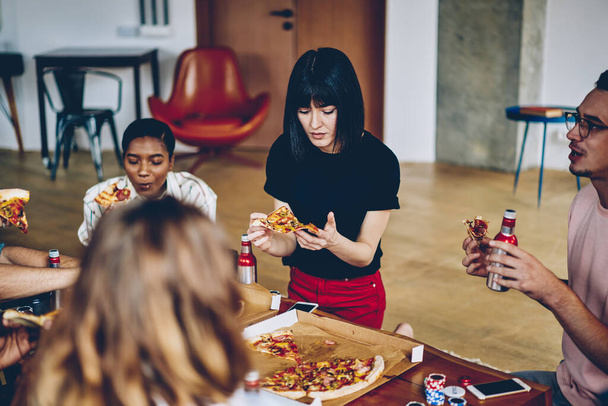Young multiracial company of friends in casual clothes eating pizza and drinking beer while playing table games during party at apartment - Fotó, kép