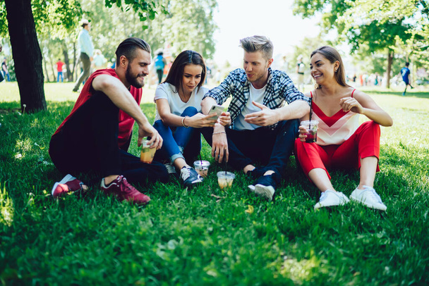 Male and female hipsters in stylish casual clothing sitting on green grass while using mobile phone smiling and gossiping together at park - Fotoğraf, Görsel