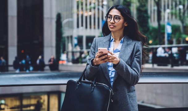 Trendy young ethnic woman in glasses and classic clothes carrying leather bag and mobile phone while walking on background of New York city - Foto, imagen