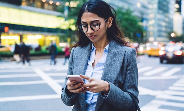 Focused serious stylish young brunette ethnic woman in round spectacles black handbag and gray coat tapping on smartphone near crosswalk on New York busy street on blurred background - Photo, image
