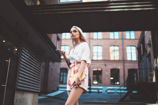 Fashionable trendy woman in light clothes holding notebooks and mobile phone in internal yard of brick building thoughtfully looking away - Foto, afbeelding