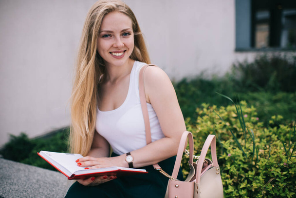 Content smiling blonde woman with pink bag with  long hair in sleeveless white shirt and green skirt reading book in red cover in street in summer - Foto, immagini