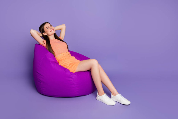Full length profile photo of funny cheerful lady sitting comfy bean bag arms behind head dreamer wear casual orange striped t-shirt jeans mini skirt isolated pastel purple color background - Φωτογραφία, εικόνα