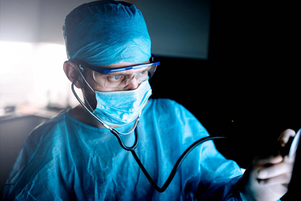 young man surgeon doctor in uniform and goggles in operating room during coronavirus pandemic. - Foto, afbeelding