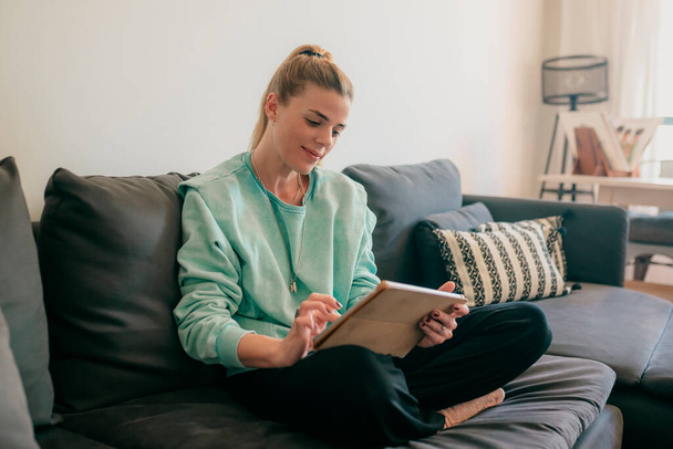 Mujer hermosa feliz viendo videos o disfrutando de contenido de entretenimiento en una tableta sentada en el sofá de la casa
. - Foto, imagen