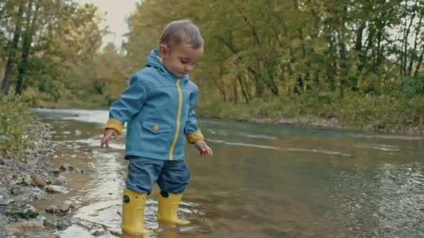 Cute baby in raincoat and rubber boots has fun by river, throws pebbles and laughs. Funny boy learns and explores natural world. Family, kids, love concept. - Footage, Video
