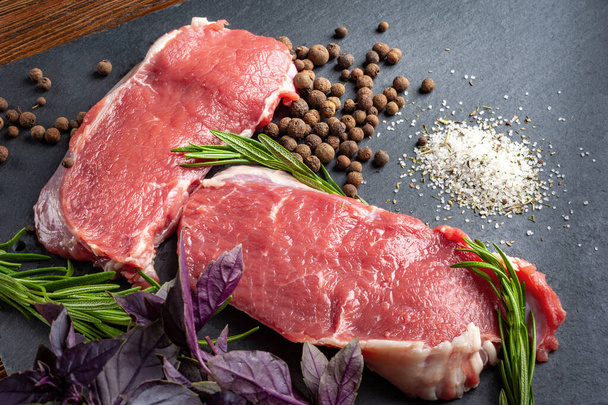 生の牛肉肉。黒スレートの石板の上に新鮮な生肉、ローズマリーと新鮮なハーブの2つのスライス。バーベキュー肉の準備とマリネ。健康的で自然食品. - 写真・画像