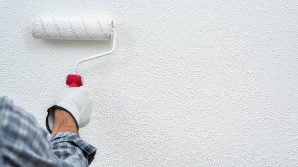 Kaukaski malarz domowy w białych kombinezonach i rękawiczkach ochronnych, malujący ścianę białą farbą za pomocą wałka. Budownictwo. Bezpieczeństwo pracy. - Zdjęcie, obraz