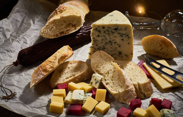 Κομμάτι τυρί με μπλε μούχλα και άλλα είδη μικρών κύβων σε φέτες σε περγαμηνή με φρέσκια μπαγκέτα και λουκάνικο, ποτήρια κρασιού. Gourmet νεκρή φύση. - Φωτογραφία, εικόνα