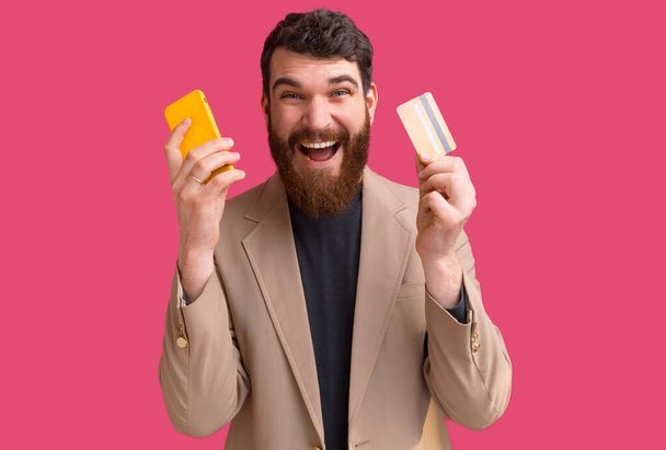 bearded man is holding a yellow phone and a card on pink background. - Foto, immagini