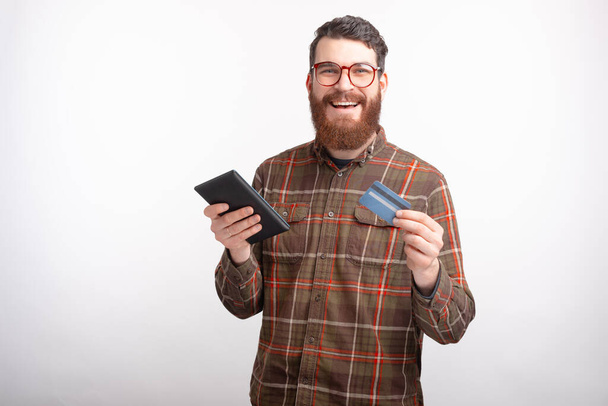 Happy γενειοφόρος άνθρωπος χαμογελά στην κάμερα, ενώ κρατώντας ένα tablet και πιστωτική ή χρεωστική κάρτα σε λευκό. - Φωτογραφία, εικόνα