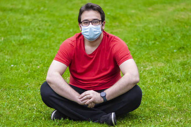 Egzersizden sonra dinlenmek için çimlerin üzerinde oturan Corona 'ya karşı önlem almak için yüz maskesi takan şişman bir adam. - Fotoğraf, Görsel