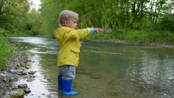 Cute baby in raincoat and rubber boots has fun by river, throws pebbles and laughs. Funny boy learns and explores natural world. Family, kids, love concept. - Footage, Video