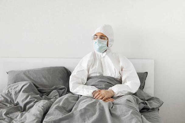 Portrait of man in protective white suit and face mask sitting in his bed in his bedroom because of covid-2019 and wait for a end of coronavirus quarantine. Self isolation. - Foto, Bild