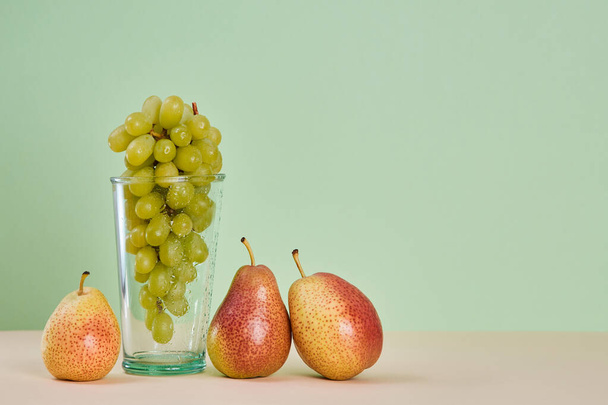 Bunch of grapes in glass. Juicy pears on beige table. Diet. Summer fruits concept. Copy space on green background - Photo, image