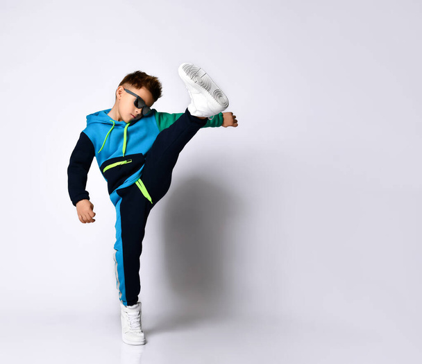 Little kid in sunglasses, colorful sport suit and sneakers. He performing a kick by his leg, posing isolated on white - Foto, Imagem