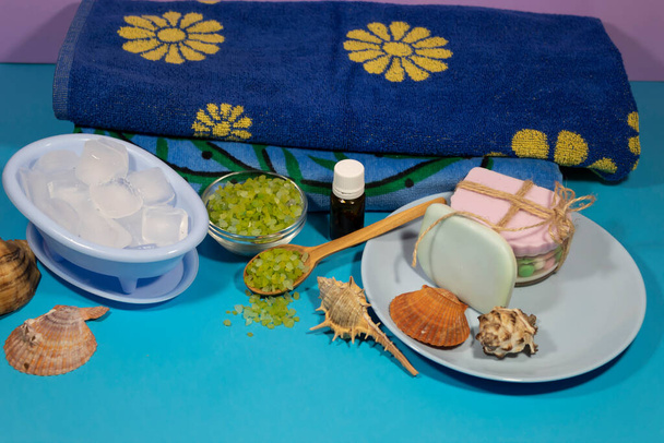 Auf blauem Hintergrund befinden sich Gegenstände für Wellnessbehandlungen - Seife, Eisstücke, Meersalz und Aromaöl. - Foto, Bild