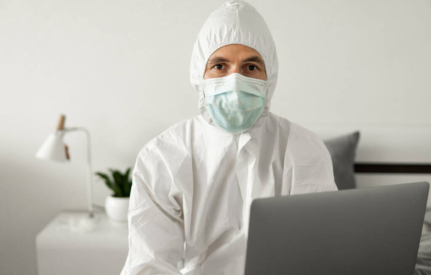 防護服を着た男性と医療用マスクは、コロナウイルスの流行のために、ラップトップ付きのベッドで自宅から働いています。パンデミック時の遠隔作業。COVID-19隔離コンセプト中に家にいます. - 写真・画像