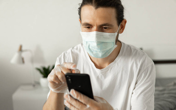 Man in medical mask is using a phone at his home sitting on a bed because of coronavirus epidemic. Remote work during pandemic. Stay home during COVID-19 quarantine concept. - Foto, Imagen