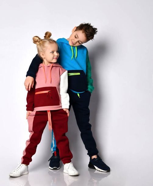 Smiling elder brother putting hand on shoulder of younger sister. Both kids in stylish track suits with cute hairdos. Portrait isolated on light grey - Foto, Bild