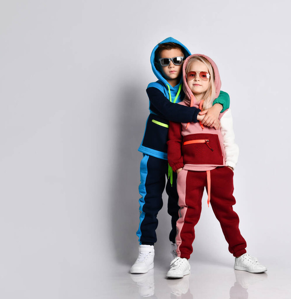 Elder brother hugging his younger sister. Stylish sporty kids in colourful tracksuits and sunglasses. Full length portrait isolated on light grey. - Photo, image