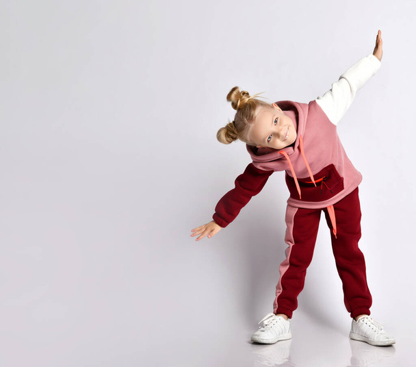 Little blonde kid with buns hairstyle, in colorful tracksuit, sneakers. Smiling, performing exercises, posing isolated on white - Фото, изображение