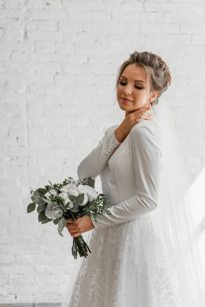 beautiful young bride with a bouquet of beautiful flowers posing against a white studio brick wall,gorgeous wedding dress of a pretty bride,fashionable wedding dress,floristry of a wedding bouquet - Photo, image