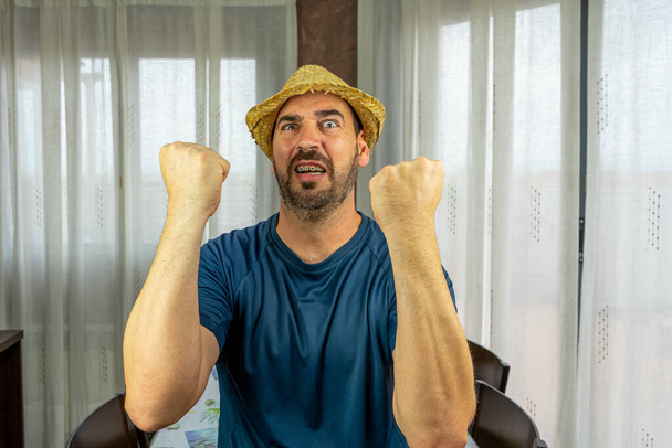 Бородатый мужчина в соломенной шляпе, одетый в темно-голубую рубашку с выражением ярости в гостиной дома. Веселая концепция
 - Фото, изображение