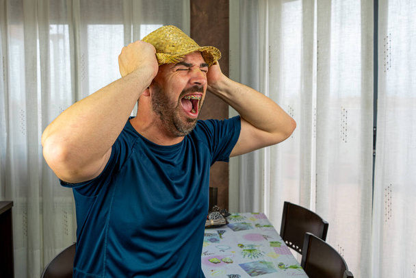 Hombre barbudo con un sombrero de paja vestido con una camisa azul oscuro con una expresión de furia en la sala de estar en casa. Concepto divertido
 - Foto, imagen