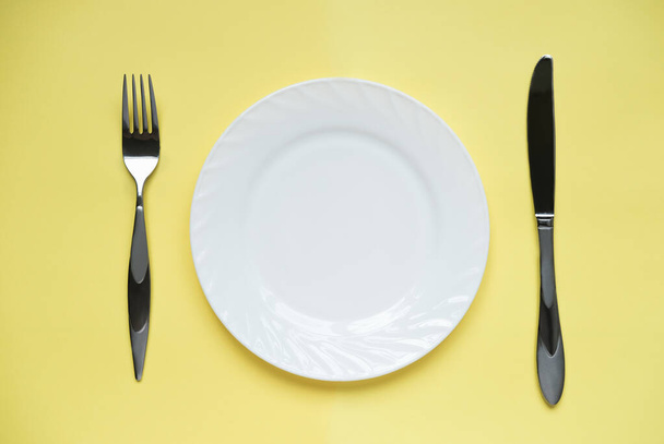 黄色の背景に白いプレートと金属製のフォークとナイフ。レストランのメニューモックアップ。食品デザインのテンプレートです。テキストと広告のためのコピースペース. - 写真・画像
