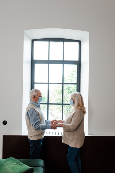 sad elderly couple in medical masks holding hands and standing near window during quarantine - Fotoğraf, Görsel